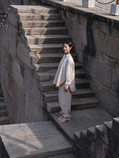 Woman wearing the Sutra Arc Stripe co-ord — a striped cotton button-down shirt with matching ruffled-hem trousers