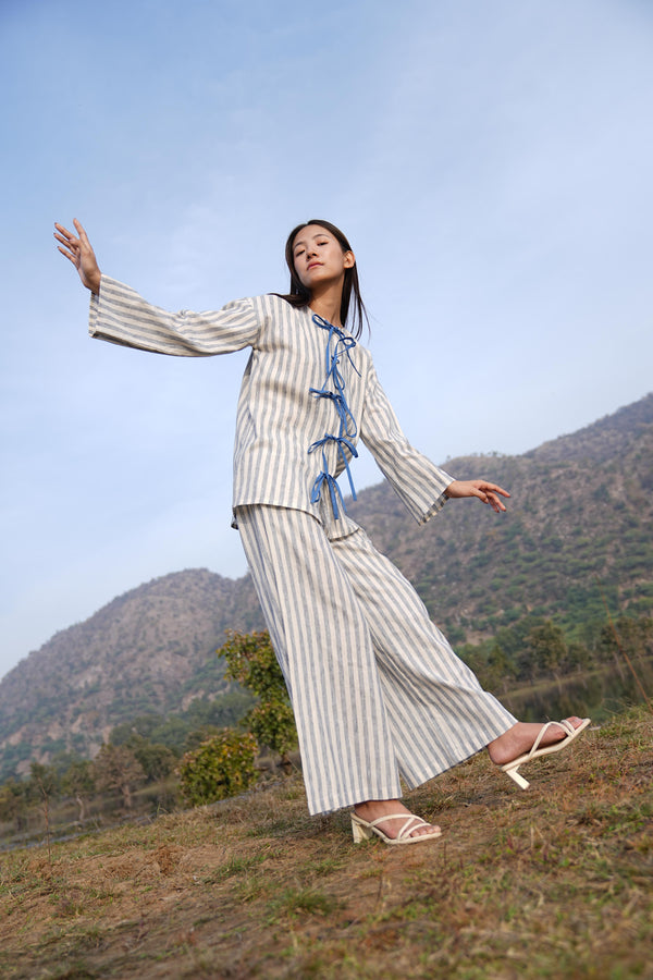 Woman wearing the Sutra Bow Stripe co-ord — an open-front striped cotton tunic tied with blue fabric ties over wide-leg trousers